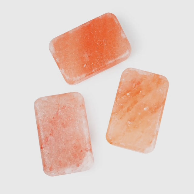 A collection of Himalayan salt body stones in various sizes, some placed on a glass dish and others scattered on a striped towel next to a potted plant, with a background of white tiles.
