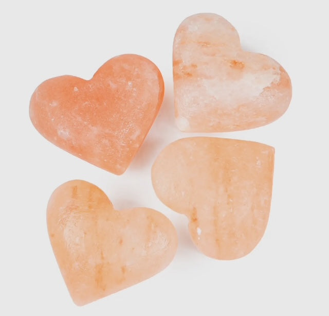 A collection of Himalayan salt body stones in various sizes, some placed on a glass dish and others scattered on a striped towel next to a potted plant, with a background of white tiles.