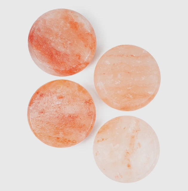 A collection of Himalayan salt body stones in various sizes, some placed on a glass dish and others scattered on a striped towel next to a potted plant, with a background of white tiles.