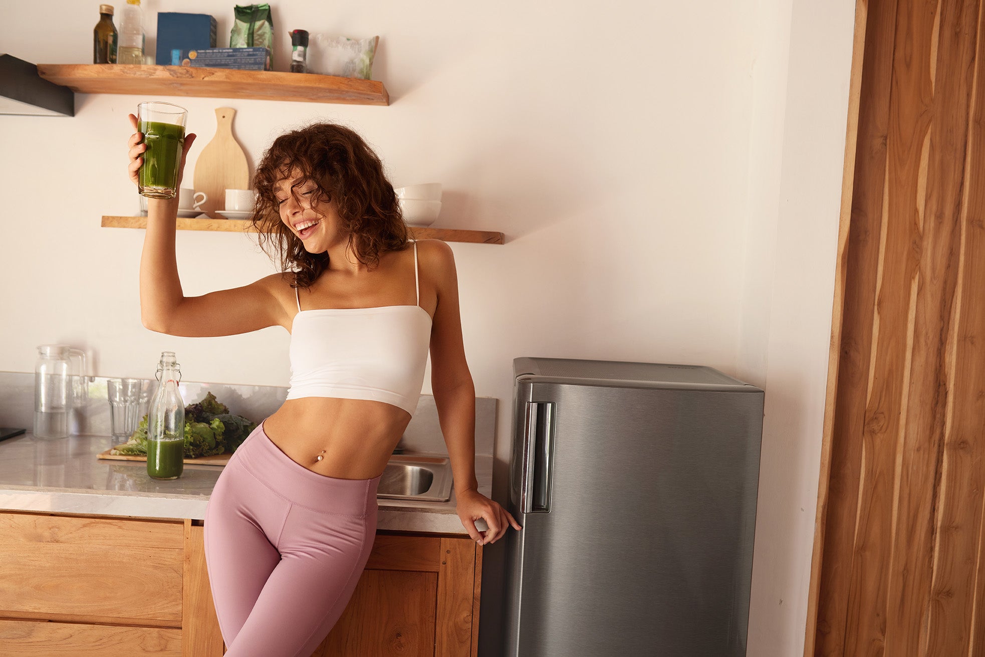 Woman in a kitchen holding a green drink, standing next to a refrigerator.